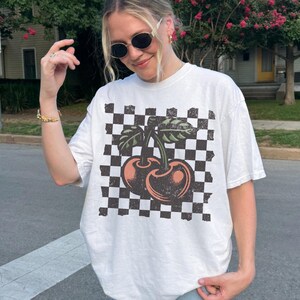 Female model wearing a white Comfort Colors checkered cherries t-shirt – vintage distressed streetwear tee with retro 90s Y2K aesthetic.