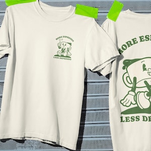 以下が含まれることがあります： 2枚のオフホワイトのTシャツ。緑色のグラフィック。片方のシャツには、「MORE ESPRESSO LESS DEPRESSO」という文字が入った漫画のコーヒーカップが描かれています。もう一方のシャツは同じデザインですが、コーヒーカップは小さく、前面にあります。