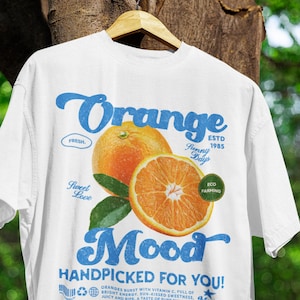 Puede incluir: Camiseta blanca con un diseño gráfico de estilo vintage que presenta una naranja con el texto "Orange Mood Handpicked for You!"  Las naranjas están llenas de vitamina C, energía brillante, dulzura bañada por el sol, jugosas y maduras, un sabor a puro sol. Más