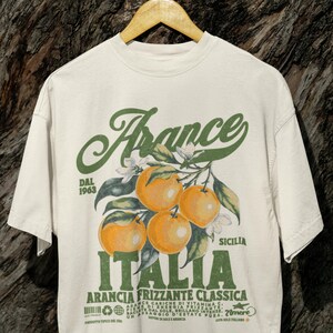 Comfort Colors® unisex T-shirt in ivory with a bold retro-style print of oranges and green vintage Italian typography. Hanging on a wooden hanger against tree bark for a rustic, nature-inspired backdrop.