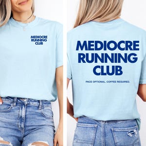 以下が含まれることがあります： 左胸にダークブルーでMEDIOCRE RUNNING CLUBの文字がプリントされ、背面にはより大きなサイズで同じ文字と、その下にPACE OPTIONAL. COFFEE REQUIRED.の文字がプリントされたライトブルーの半袖Tシャツ。