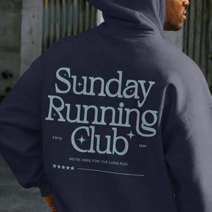 May include: A dark navy blue hooded sweatshirt featuring the white text Sunday Running Club on the back. The design includes small star icons, the text ESTD 1991, the phrase WE'RE HERE FOR THE LONG RUN, and a row of five stars above a horizontal line.