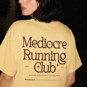 May include: A light yellow t-shirt with the words "Mediocre Running Club" in brown lettering. The shirt also features the text "ESTD 1991" and "ENJOYING EVERY STEP AT OUR OWN PACE".