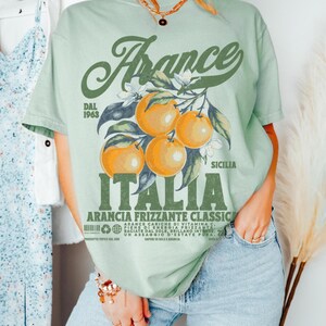 May include: Sage green t-shirt with a vintage-style graphic featuring oranges, flowers, and the words "France" and "Italia." The design includes text that reads "Arancia Frizzante Classic" and "Dal 1963." The model is wearing an XL size.