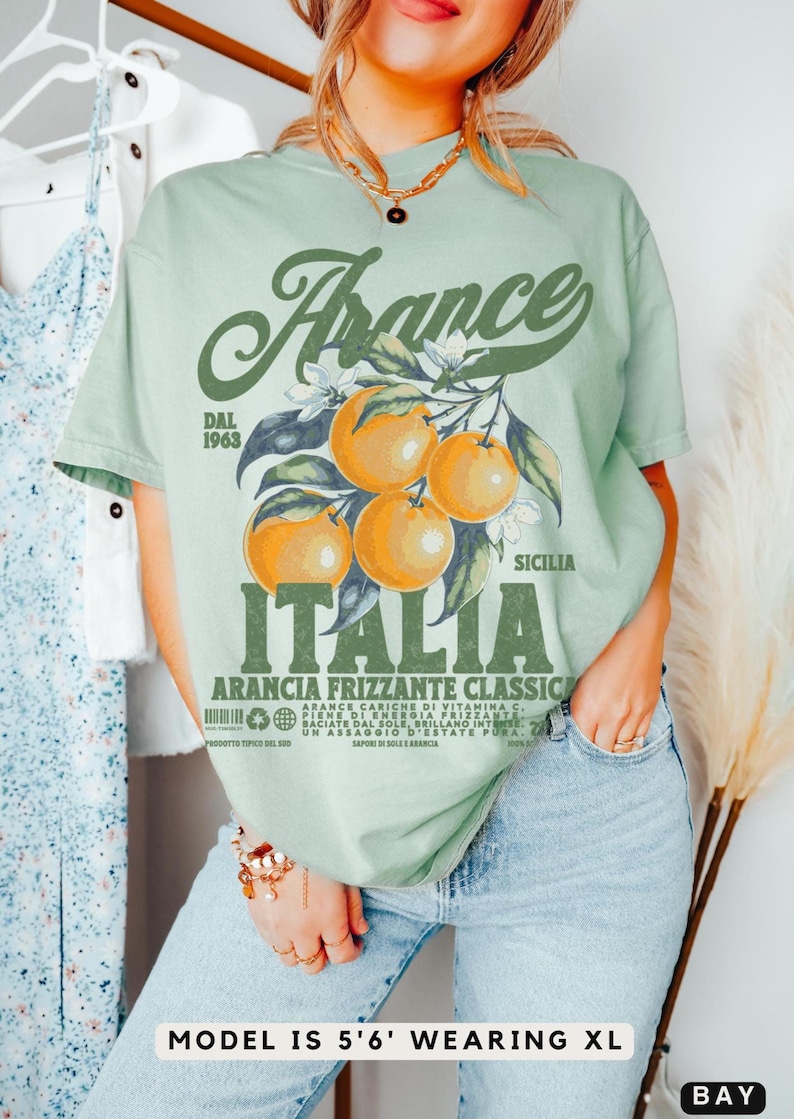 May include: Sage green t-shirt with a vintage-style graphic featuring oranges, flowers, and the words "France" and "Italia." The design includes text that reads "Arancia Frizzante Classic" and "Dal 1963." The model is wearing an XL size.