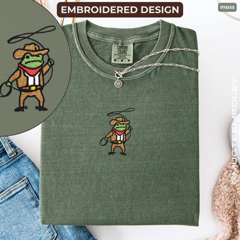 Comfort Colors Moss embroidered cowboy frog tee flat lay with ‘embroidered design’ label and zoomed detail; stitched cowboy frog with lasso shown close up.