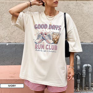 May include: An ivory short-sleeve t-shirt featuring a graphic print with the text "GOOD DAYS RUN CLUB" and "Fueled by coffee & Endorphins". The design includes illustrations of a pair of athletic sneakers and a glass of iced coffee.