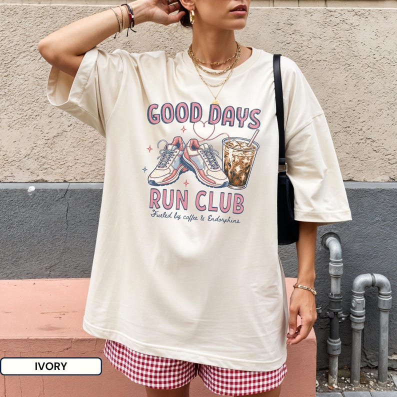 May include: An ivory short-sleeve t-shirt featuring a graphic print with the text "GOOD DAYS RUN CLUB" and "Fueled by coffee & Endorphins". The design includes illustrations of a pair of athletic sneakers and a glass of iced coffee.