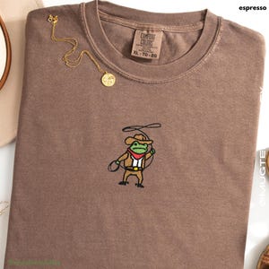 Comfort Colors Espresso embroidered cowboy frog t-shirt folded flat lay with necklace; tiny western frog embroidery centered.