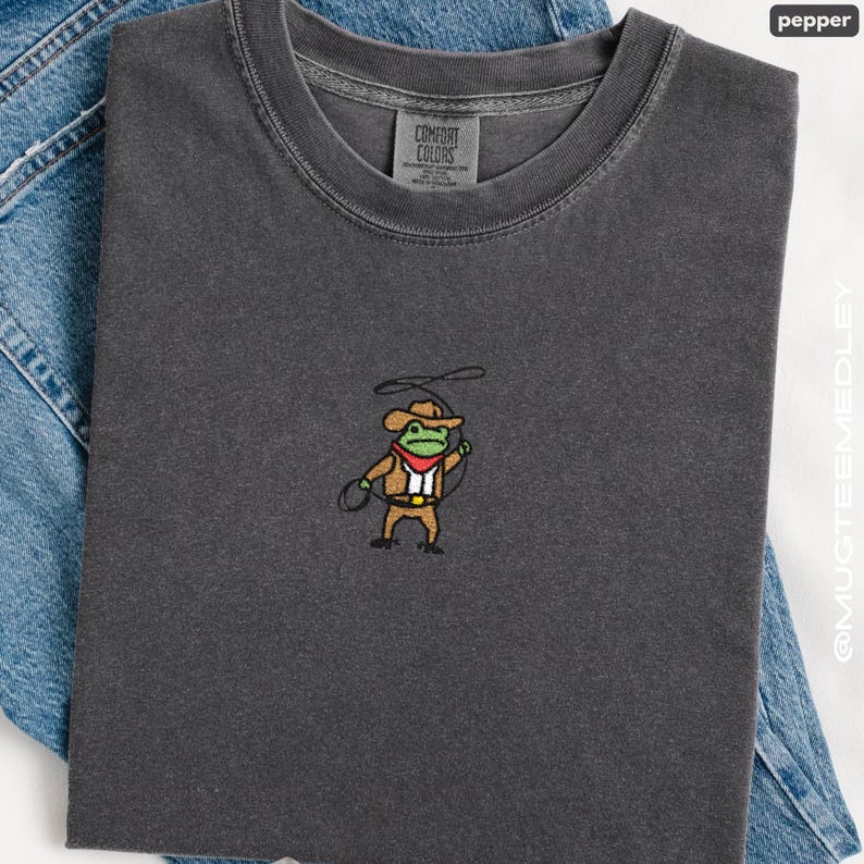 Comfort Colors Pepper embroidered cowboy frog tee folded on denim; minimal chest embroidery of a cute cowboy frog with lasso