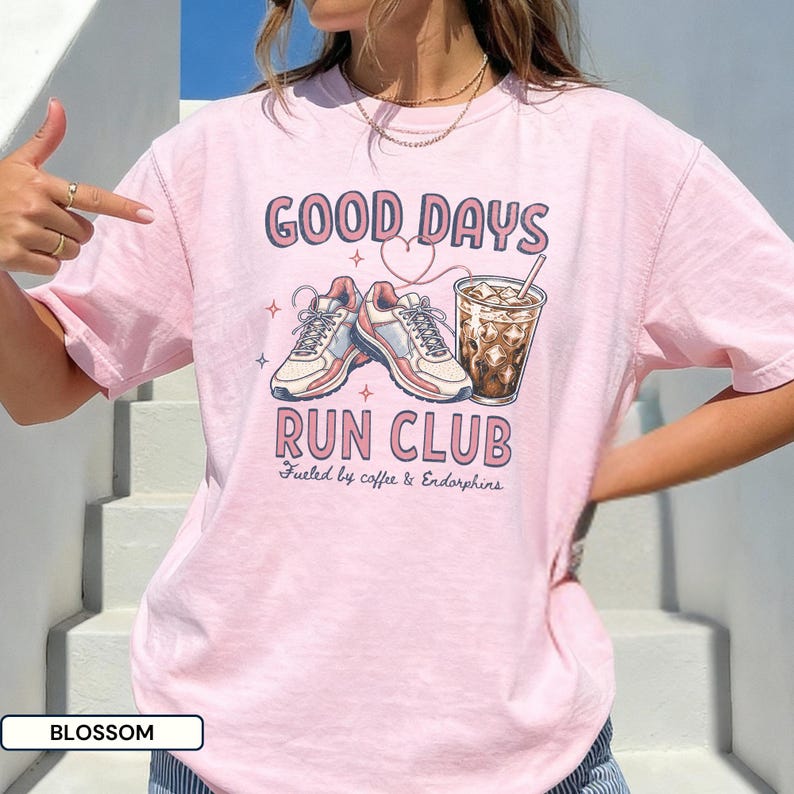 May include: A light pink t-shirt featuring a graphic print with the text GOOD DAYS RUN CLUB and Fueled by coffee & Endorphins. The design includes a pair of athletic sneakers, a heart outline, and a glass of iced coffee with a straw.