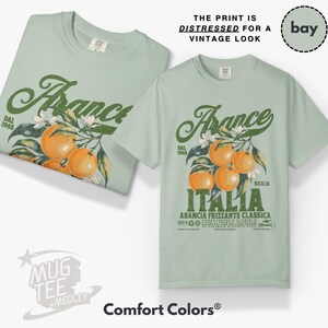 May include: Two light green t-shirts with a vintage-style graphic. The graphic features the word "Arance" in a distressed green font, with orange fruit and the word "Italia" below. The text "THE PRINT IS DISTRESSED FOR A VINTAGE LOOK" is above.