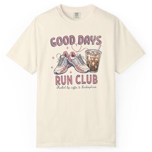 May include: A cream-colored short-sleeve t-shirt featuring a graphic of running shoes and an iced coffee. The text reads GOOD DAYS RUN CLUB and Fueled by coffee & Endorphins. The design includes small star and heart outlines in pink and red.