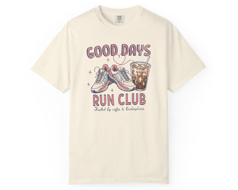 May include: A cream-colored short-sleeve t-shirt featuring a graphic of running shoes and an iced coffee. The text reads GOOD DAYS RUN CLUB and Fueled by coffee & Endorphins. The design includes small star and heart outlines in pink and red.