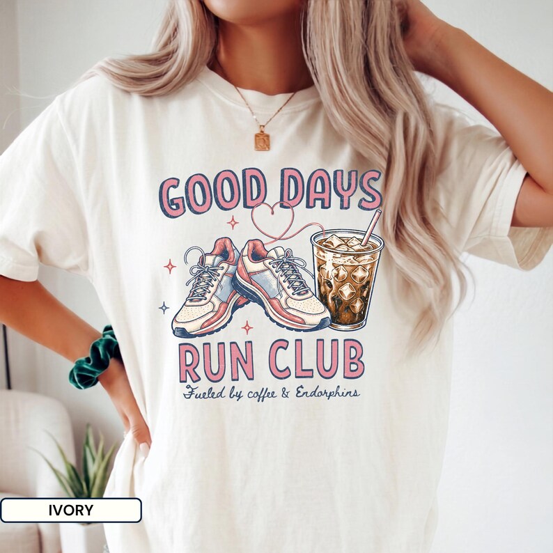 May include: An ivory t-shirt featuring a graphic print with the text GOOD DAYS RUN CLUB and Fueled by coffee & Endorphins. The design includes a pair of athletic sneakers, a heart outline, and a glass of iced coffee with a straw in pink and blue.