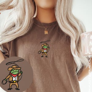 Close-up of model wearing Comfort Colors Espresso embroidered cowboy frog tee; small chest embroidery visible, cozy lifestyle photo with mug