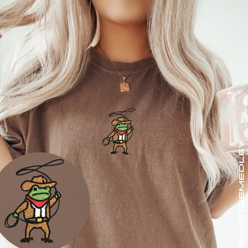 Close-up of model wearing Comfort Colors Espresso embroidered cowboy frog tee; small chest embroidery visible, cozy lifestyle photo with mug