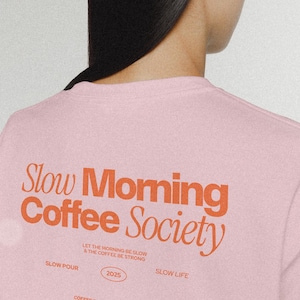 Op de afbeelding: Roze T-shirt met de tekst "Slow Morning Coffee Society" in oranje. De tekst "Let the morning be slow & the coffee be strong" staat onder de hoofdtekst. De tekst "Slow Pour" en "Slow Life" staan aan weerszijden van het jaar "2025". De tekst "Coffee first, everything later" staat onder het jaar.