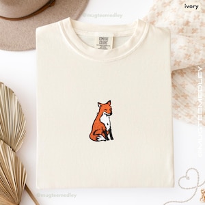 Peut inclure: T-shirt Comfort Colors de couleur ivoire avec un petit renard brodé en orange et blanc. Le renard est assis, les yeux fermés. Le t-shirt est plié proprement. Le mot "ivoire" est visible en haut à droite.