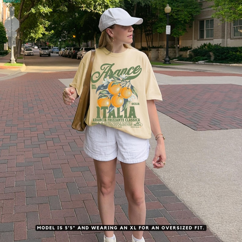 Woman standing outdoors wearing a pale yellow Comfort Colors t-shirt with a vintage Italian orange soda graphic. The design features bold green and orange typography with illustrations of oranges and the words "Arancia Frizzante Classica" and "Italia Sicilia." The model is 5'5" and wearing an XL for an oversized fit. Urban background with trees and brick road.