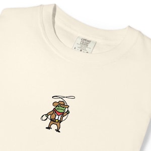 May include: Cream-colored sweatshirt with a small embroidered design of a frog dressed as a cowboy, holding a lasso. The frog wears a brown hat, jacket, and pants, with a red neckerchief. The tag reads "Comfort Colors".