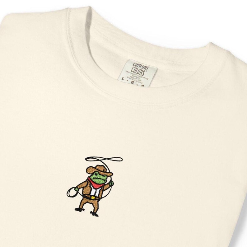 May include: Cream-colored sweatshirt with a small embroidered design of a frog dressed as a cowboy, holding a lasso. The frog wears a brown hat, jacket, and pants, with a red neckerchief. The tag reads "Comfort Colors".