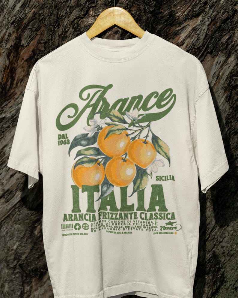 Comfort Colors® unisex T-shirt in ivory with a bold retro-style print of oranges and green vintage Italian typography. Hanging on a wooden hanger against tree bark for a rustic, nature-inspired backdrop.