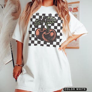 May include: White oversized t-shirt with a black and white checkered pattern and a vintage-style cherry graphic. The model is 5'6" (168 cm) and usually wears a size M.  The shirt is available in sizes up to XL.