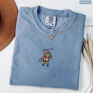 Comfort Colors Blue Jean embroidered cowboy frog t-shirt folded flat lay with necklace; small stitched frog in cowboy hat and lasso centered on chest.