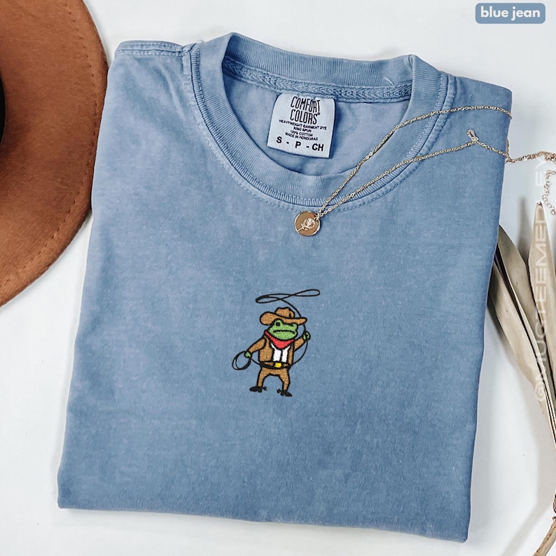 Comfort Colors Blue Jean embroidered cowboy frog t-shirt folded flat lay with necklace; small stitched frog in cowboy hat and lasso centered on chest.