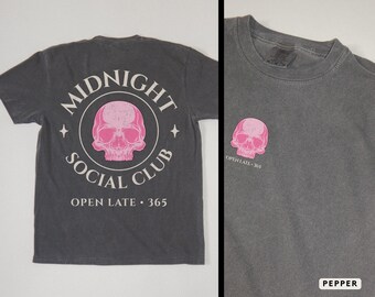 Może przedstawiać: Ciemnoszara koszulka z różowym nadrukiem czaszki. Tył koszulki zdobi okrągły wzór z napisami "MIDNIGHT SOCIAL CLUB" i "OPEN LATE 365". Mniejszy nadruk czaszki znajduje się z przodu.