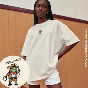 May include: Cream-colored t-shirt with a small embroidered frog wearing a cowboy hat and holding a lasso. The shirt is paired with white shorts. The image includes a close-up of the frog design.