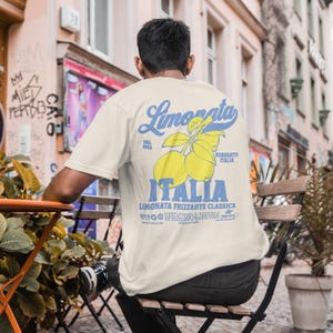 May include: Cream-colored t-shirt with a blue and yellow graphic on the back. The graphic features the word "Limonata" in script, lemons, and the word "Italia". The shirt is being worn outdoors.
