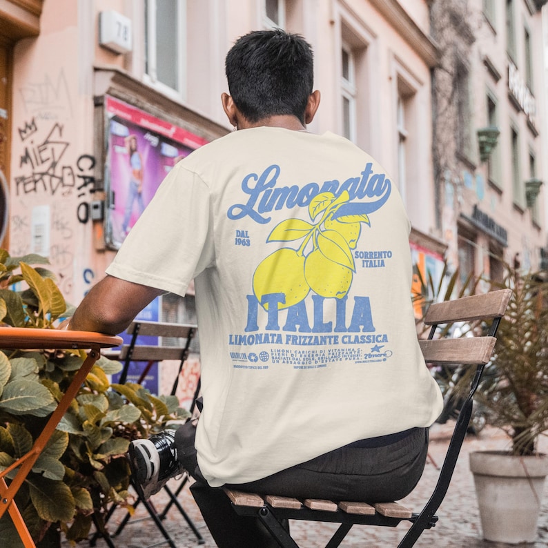 May include: Cream-colored t-shirt with a blue and yellow graphic on the back. The graphic features the word "Limonata" in script, lemons, and the word "Italia". The shirt is being worn outdoors.