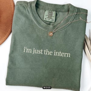 May include: A green t-shirt with the text "i'm just the intern" printed in white on the front. The shirt has a tag on the front that says "COMFORT COLORS".