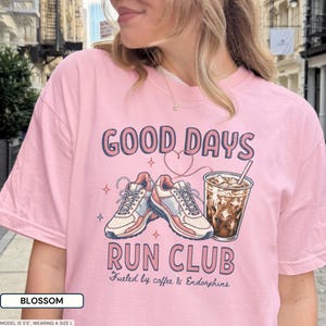 May include: A light pink t-shirt featuring a graphic print with the text GOOD DAYS RUN CLUB and Fueled by coffee & Endorphins. The design includes a pair of athletic sneakers, a heart outline, and a plastic cup filled with iced coffee.