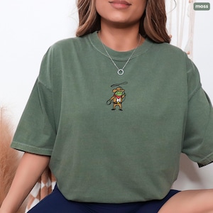 Model wearing Comfort Colors Moss embroidered cowboy frog t-shirt; relaxed oversized fit with small cowboy frog embroidery on chest.