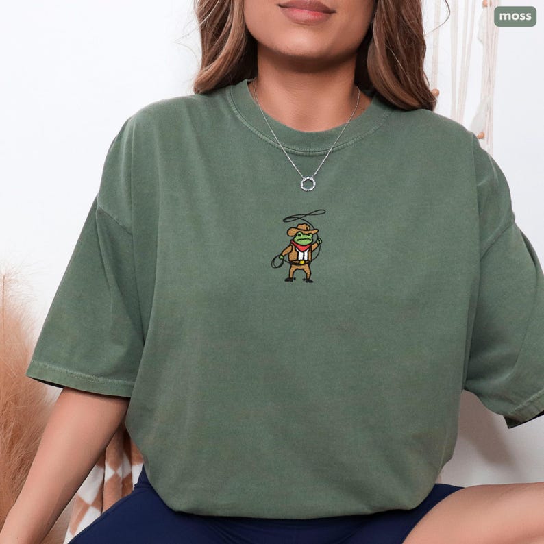 Model wearing Comfort Colors Moss embroidered cowboy frog t-shirt; relaxed oversized fit with small cowboy frog embroidery on chest.