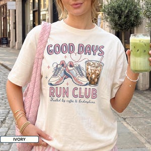 May include: An ivory t-shirt featuring a graphic print of a pair of running shoes and an iced coffee. The text reads GOOD DAYS RUN CLUB and Fueled by coffee & Endorphins. The design includes a heart outline connecting the shoes and drink.