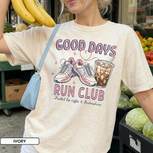 May include: An ivory short-sleeve t-shirt featuring a graphic print with the text GOOD DAYS RUN CLUB and Fueled by coffee & Endorphins. The design includes two athletic sneakers, a heart outline, and a glass of iced coffee with a straw.