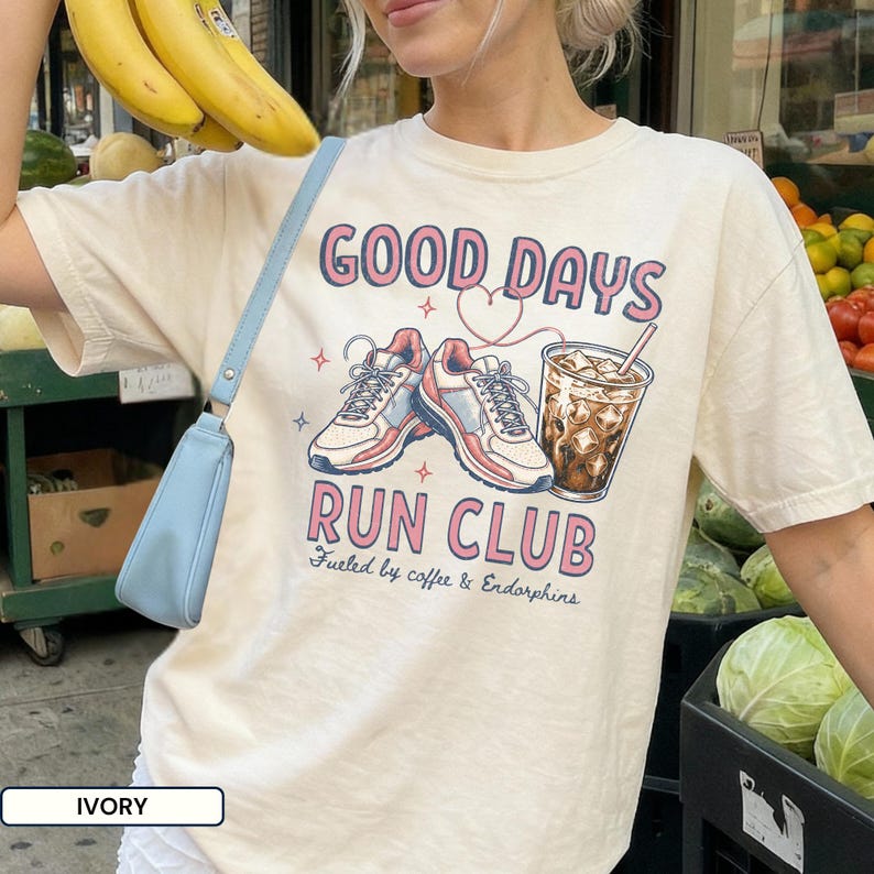 May include: An ivory short-sleeve t-shirt featuring a graphic print with the text GOOD DAYS RUN CLUB and Fueled by coffee & Endorphins. The design includes two athletic sneakers, a heart outline, and a glass of iced coffee with a straw.