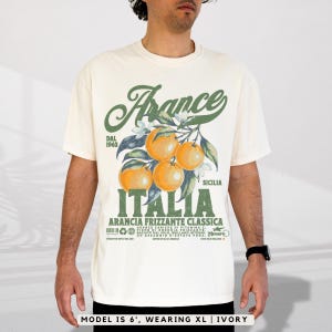 May include: Ivory-colored t-shirt featuring a graphic of oranges on a branch with the text "France Italia" in green. The shirt has text that reads "Arancia Frizzante Classica" and "Dal 1963". The model is wearing an XL.