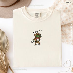Comfort Colors Ivory embroidered cowboy frog t-shirt folded flat lay with soft neutral props; tiny cowboy frog embroidery on front.