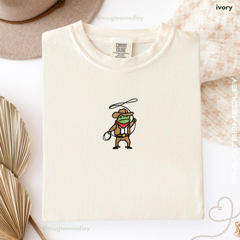Comfort Colors Ivory embroidered cowboy frog t-shirt folded flat lay with soft neutral props; tiny cowboy frog embroidery on front.