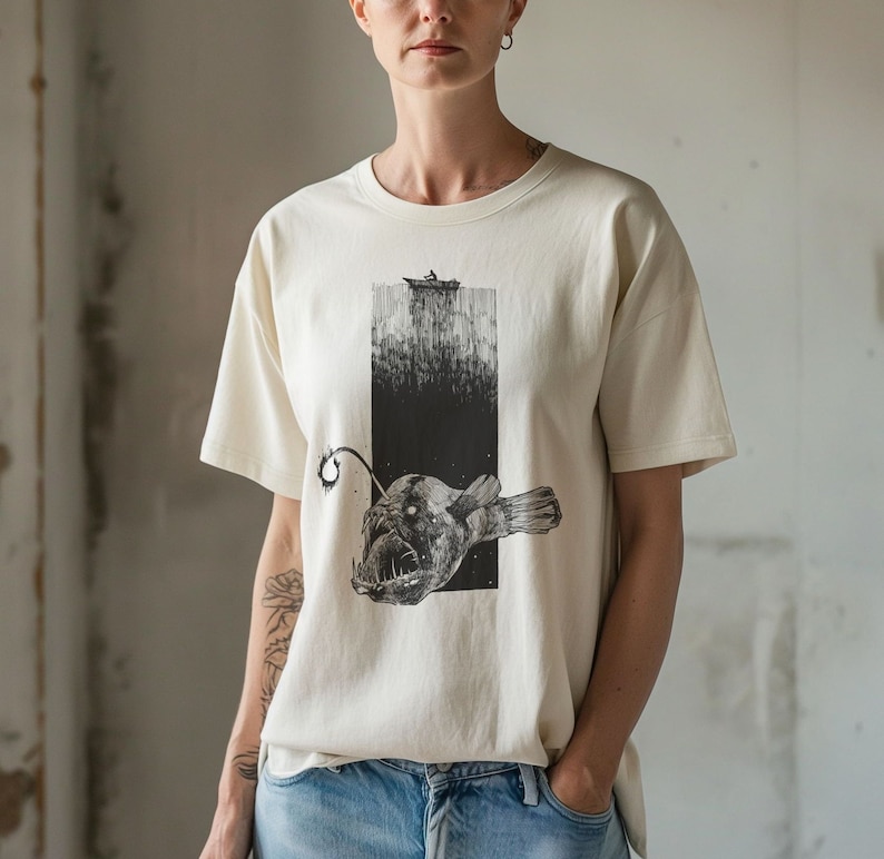 Hand-drawn anglerfish tee featuring a deep ocean scene with a boat and fisherman on the surface. Perfect for nature lovers, fans of retro 90s and 2000s style, and grunge fashion enthusiasts. Fish Shirt.