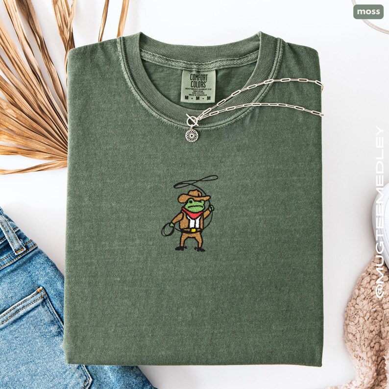Comfort Colors Moss embroidered cowboy frog tee folded with denim and neutral props; small chest embroidery of frog in cowboy outfit