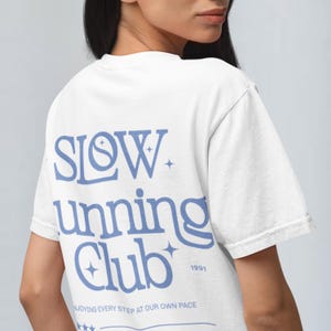 May include: White t-shirt with a blue graphic that says "SLOW running club 1991" and "ENJOYING EVERY STEP AT OUR OWN PACE".