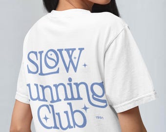 以下が含まれることがあります： 「SLOW running club 1991」と「ENJOYING EVERY STEP AT OUR OWN PACE」と書かれた青いグラフィックがプリントされた白いTシャツです。