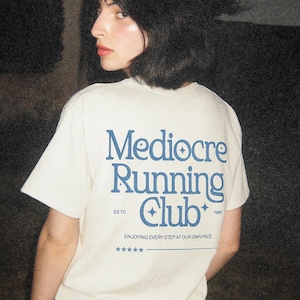 May include: A cream-colored t-shirt featuring blue text on the back that reads Mediocre Running Club, ESTD 1991, and ENJOYING EVERY STEP AT OUR OWN PACE, accompanied by small star icons and a line of five stars at the bottom of the graphic design.