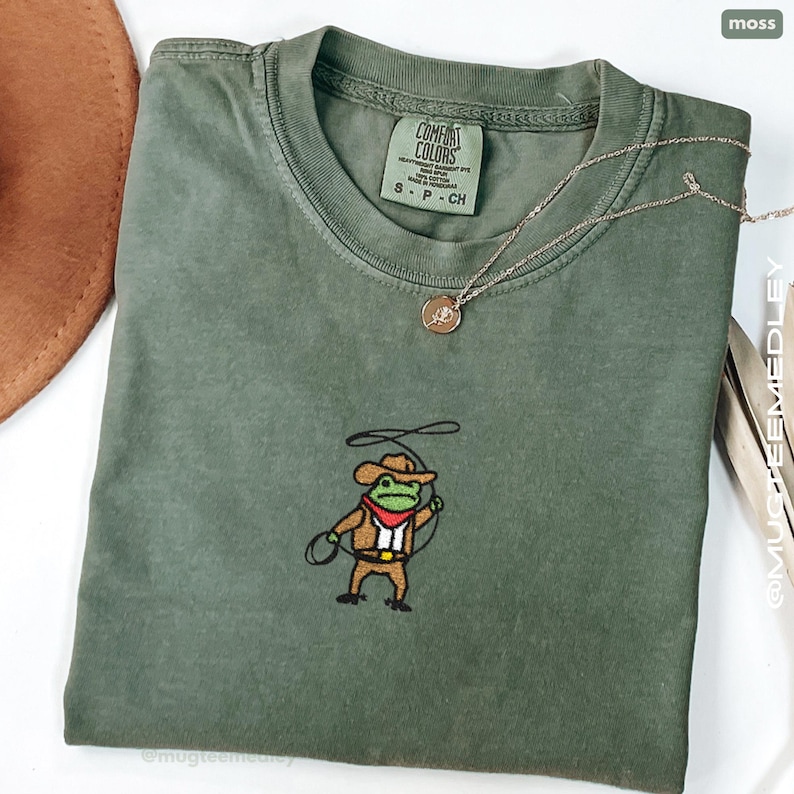 May include: Moss green t-shirt with a cowboy frog embroidery. The shirt has a crew neck and short sleeves. A gold necklace with a pendant rests on the shirt. The tag reads "Comfort Colors".